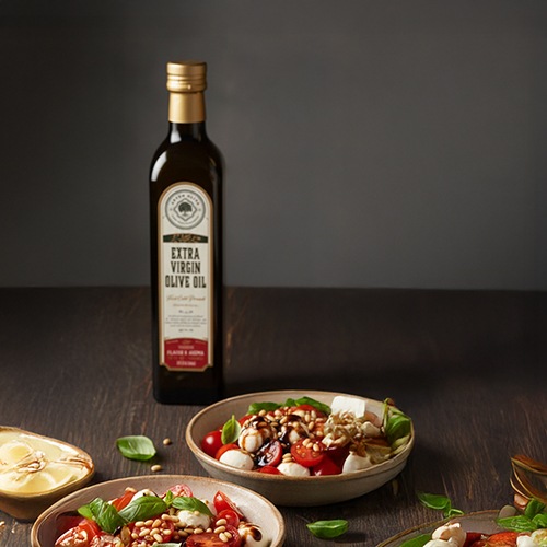 500 ml extra virgin olive oil glass bottle with gold cap standing on wooden table next to Mediterranean salad plates with tomatoes, cheese, basil, and pine nuts.