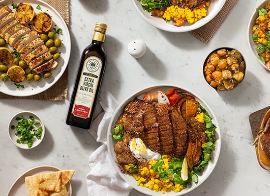 Top-down Mediterranean meal on marble table with grilled meats, olives, vegetable sides, flatbread, and a bottle of extra virgin olive oil.