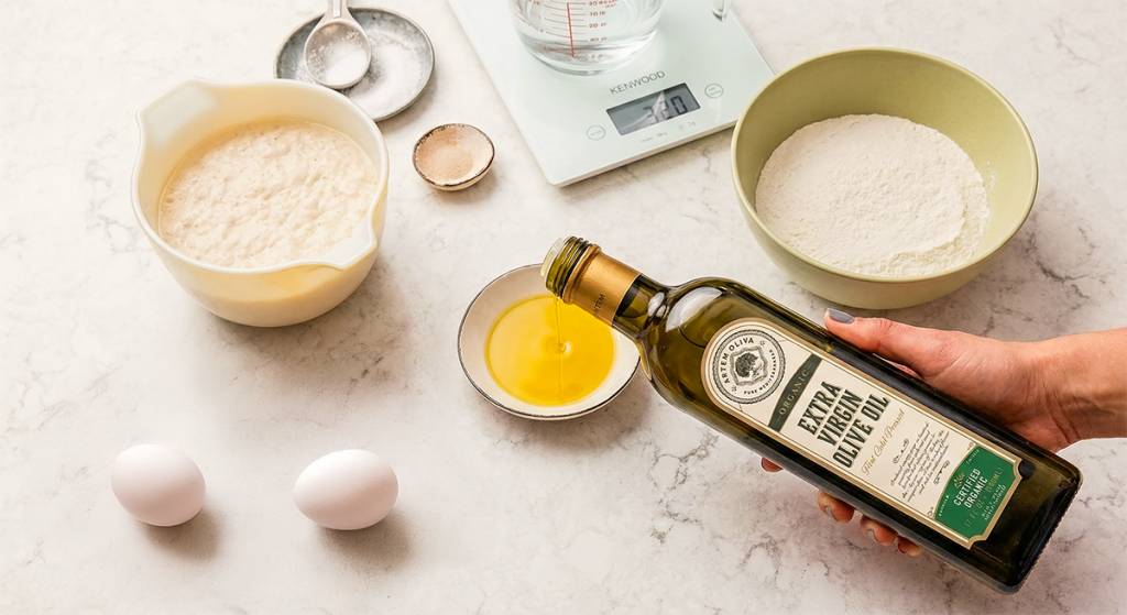 Extra virgin olive oil being poured from a glass bottle into a small bowl next to flour, eggs, and dough starter on a marble kitchen counter.