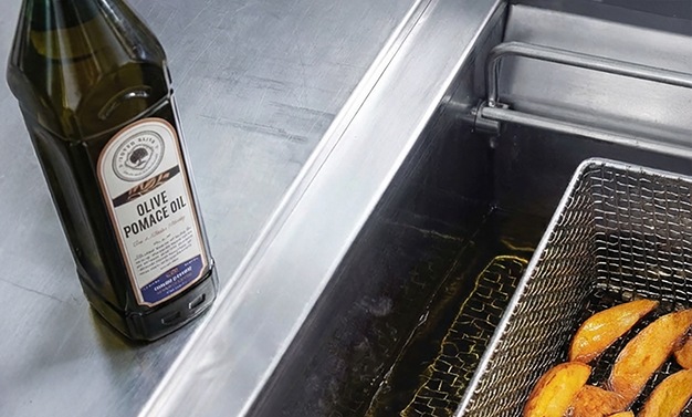 Artem Oliva olive pomace oil bottle standing next to a stainless steel deep fryer with potato wedges frying in hot oil in a commercial kitchen.
